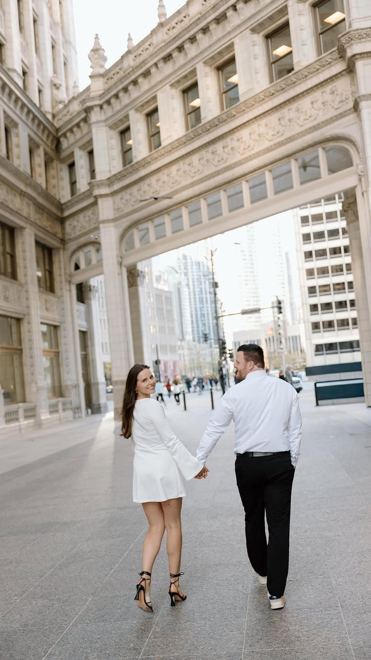 Couple holding hands and walking through a city plaza with ornate stone architecture.