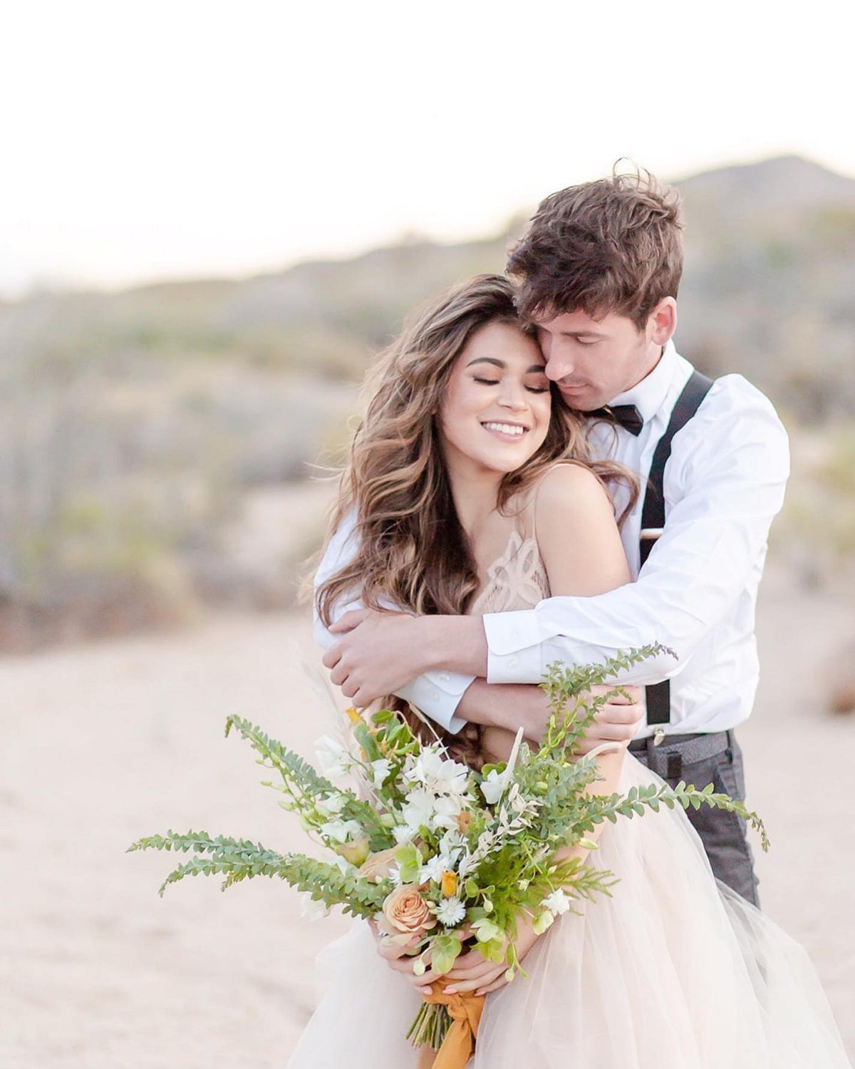 Couple embracing in a sandy landscape, woman holding a wildflower bouquet.