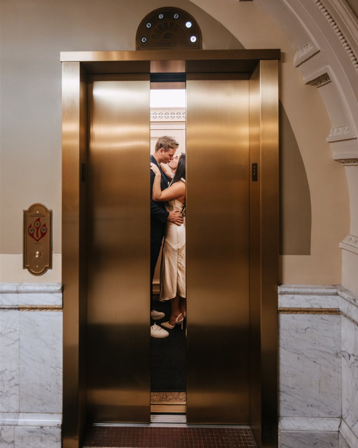 Couple kissing inside a gold elevator framed by open doors in a marble lobby