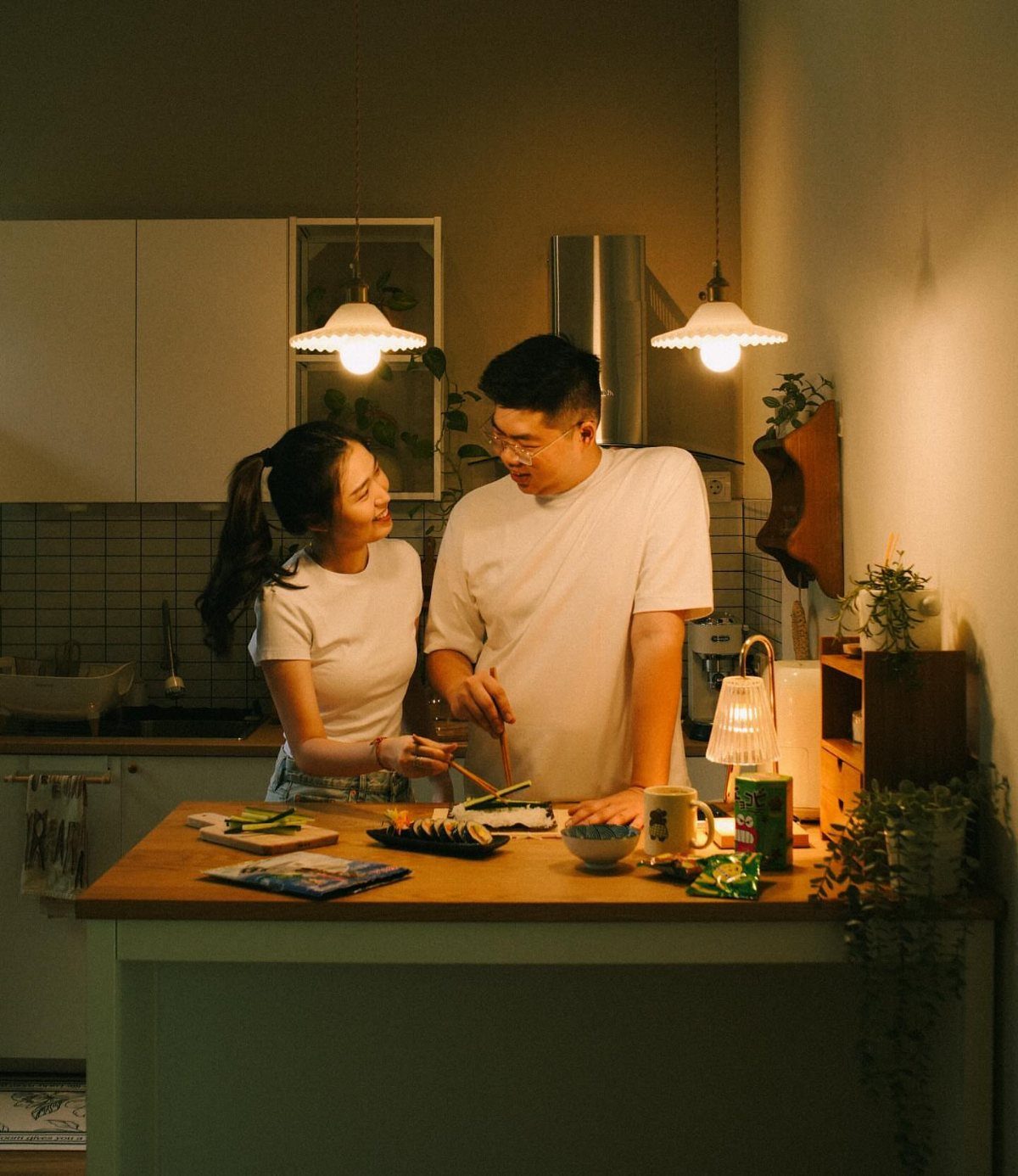 Couple in white t-shirts smiling while preparing food together in a cozy kitchen.