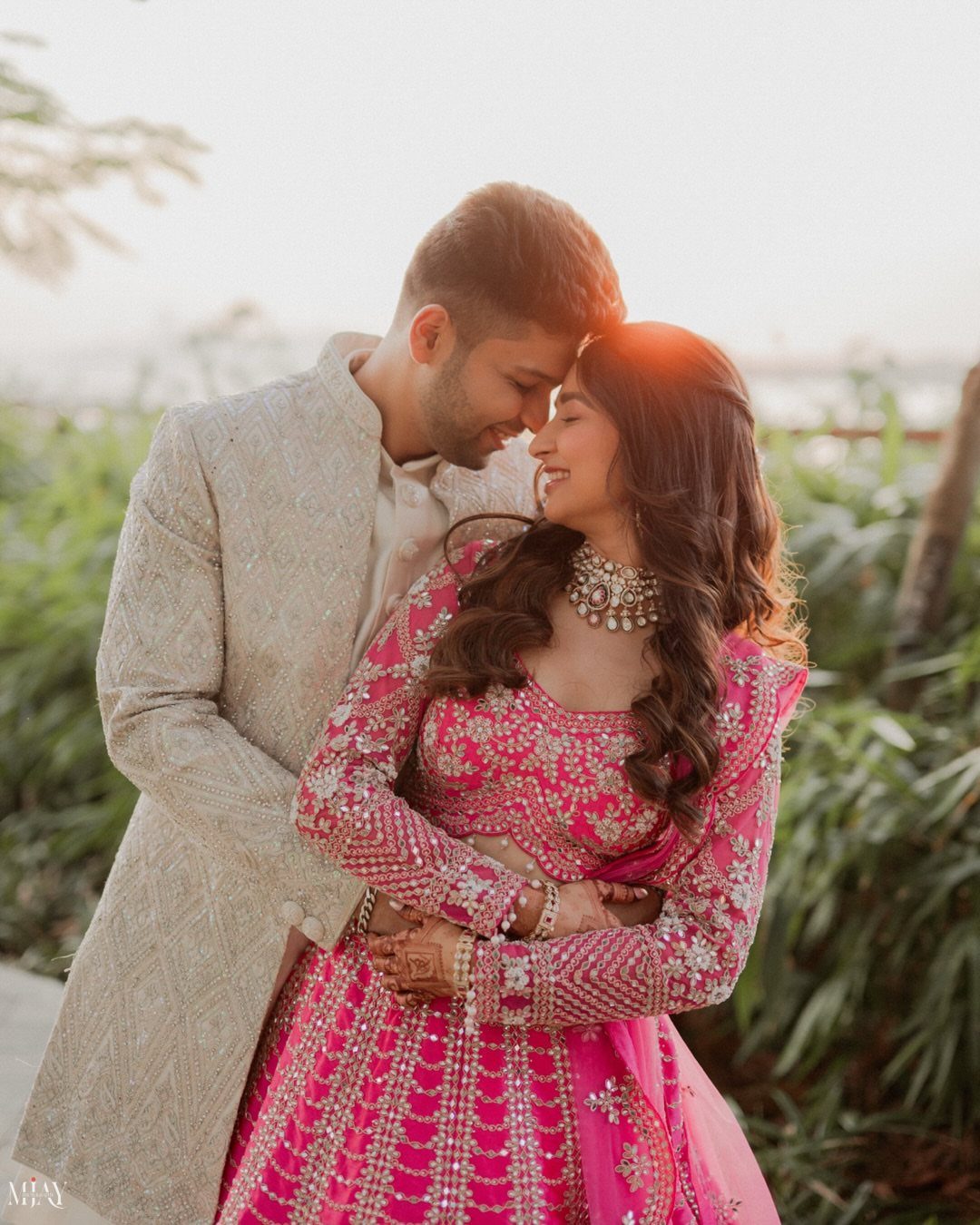 Couple in traditional Indian wedding attire touching foreheads and smiling in a garden.