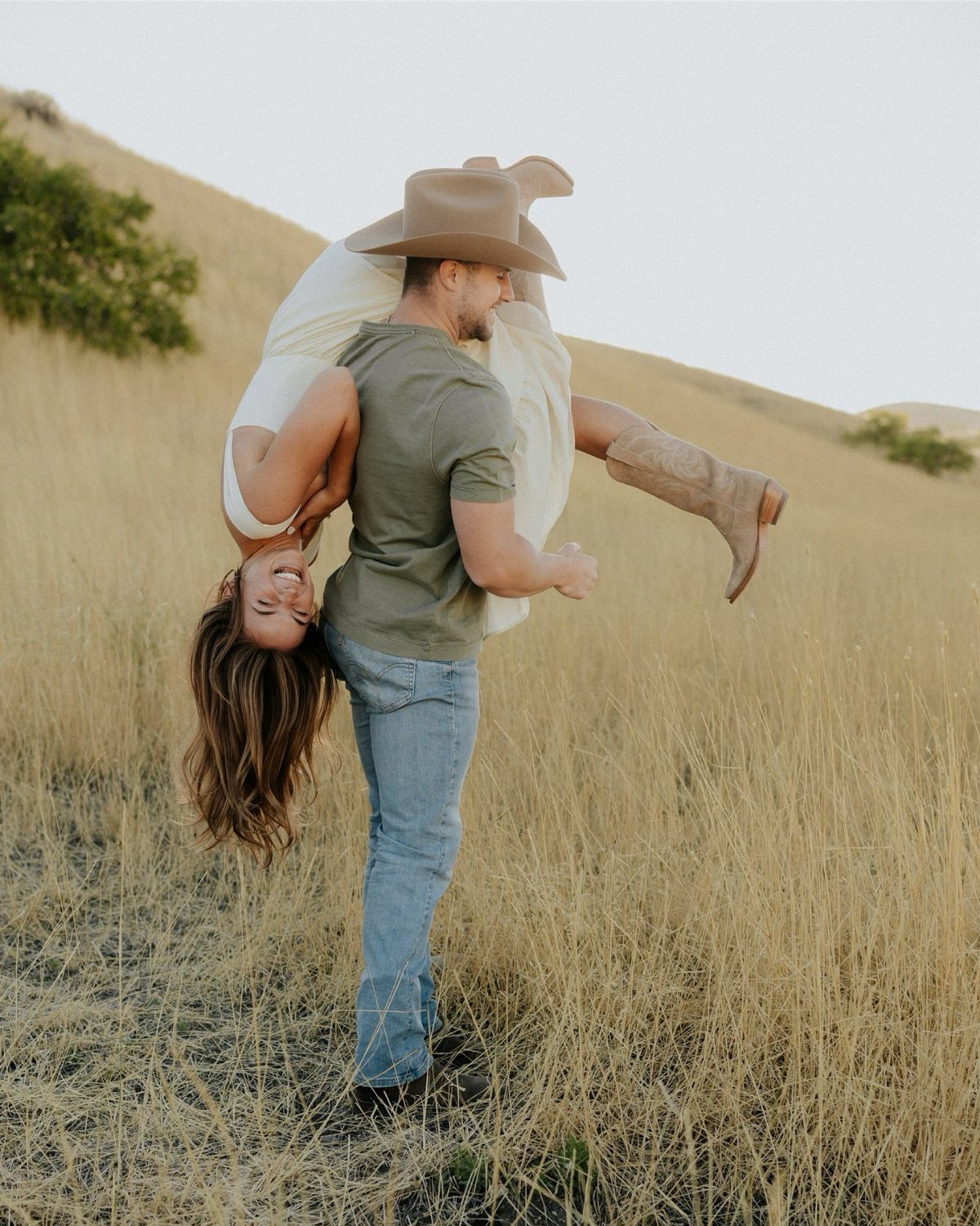 Man in cowboy hat carrying laughing woman upside down in a grassy field