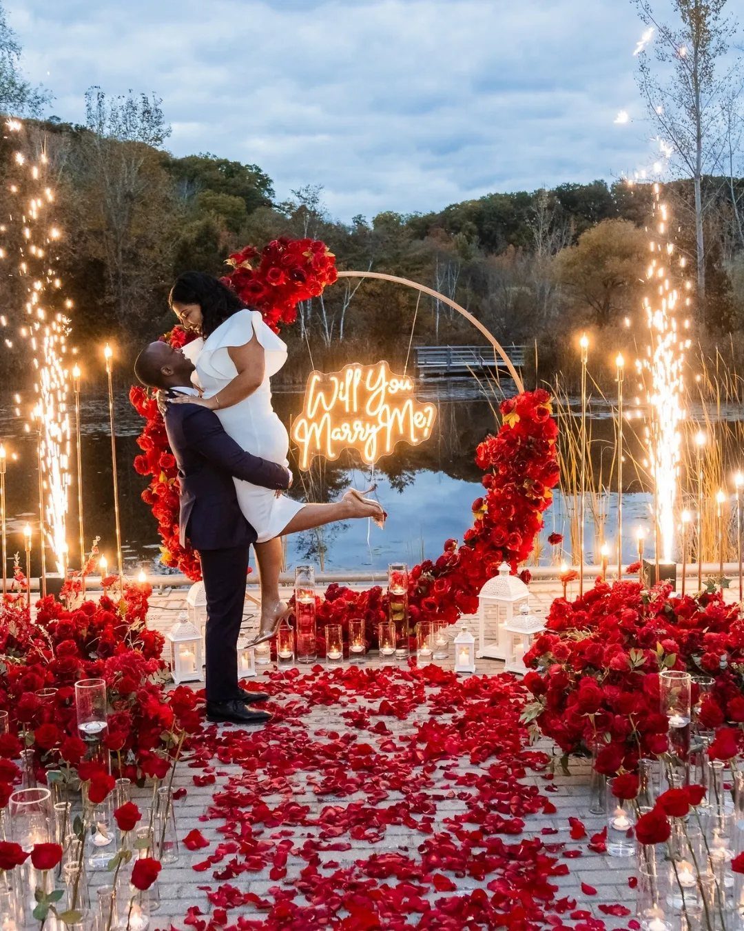 Couple embracing amidst red roses and sparks by a lake during a proposal.