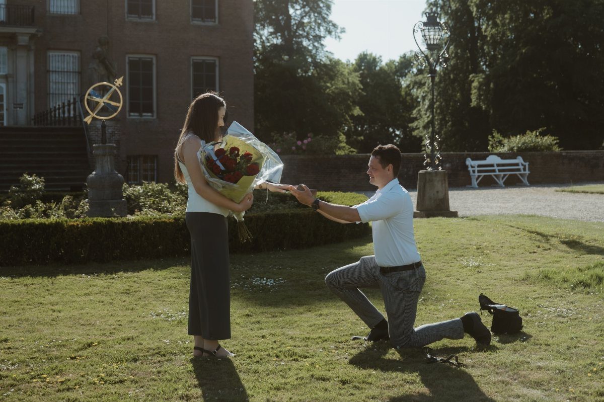 Man kneeling on a lawn proposing to a woman holding red roses.