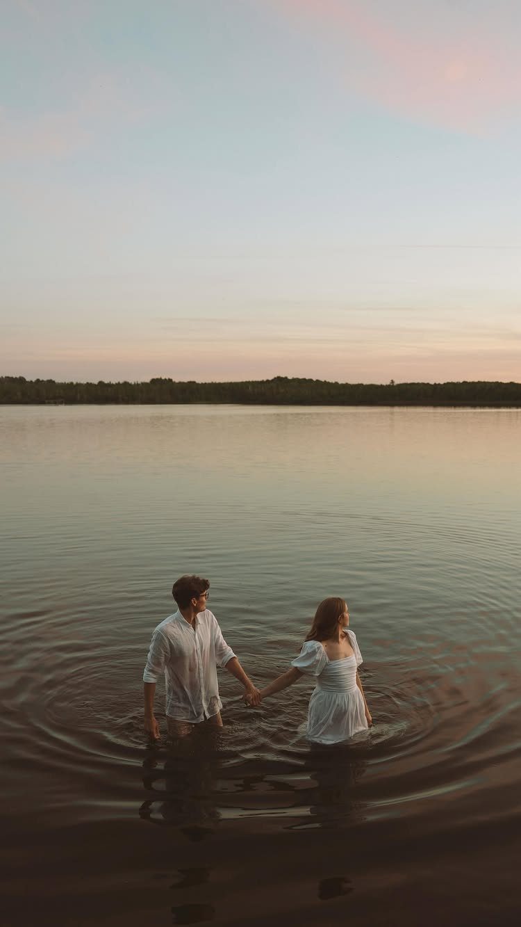 Couple in white clothing holding hands while standing in a calm lake at sunset.