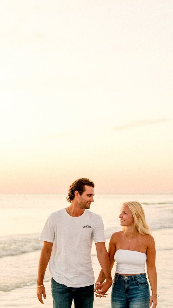 Couple walking hand-in-hand on a beach at sunset, smiling at each other.