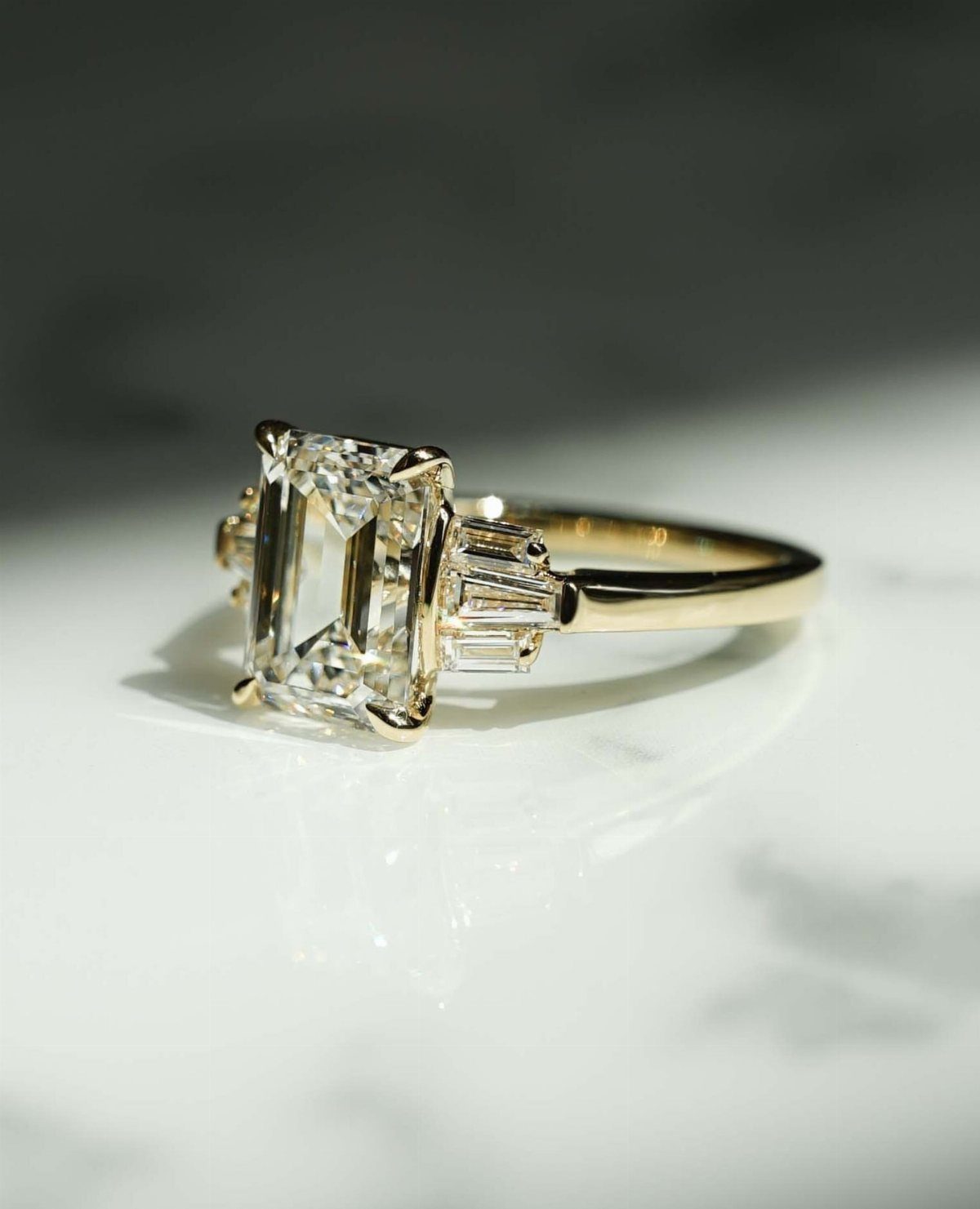 Gold engagement ring with emerald-cut diamond and baguette side stones on white marble.