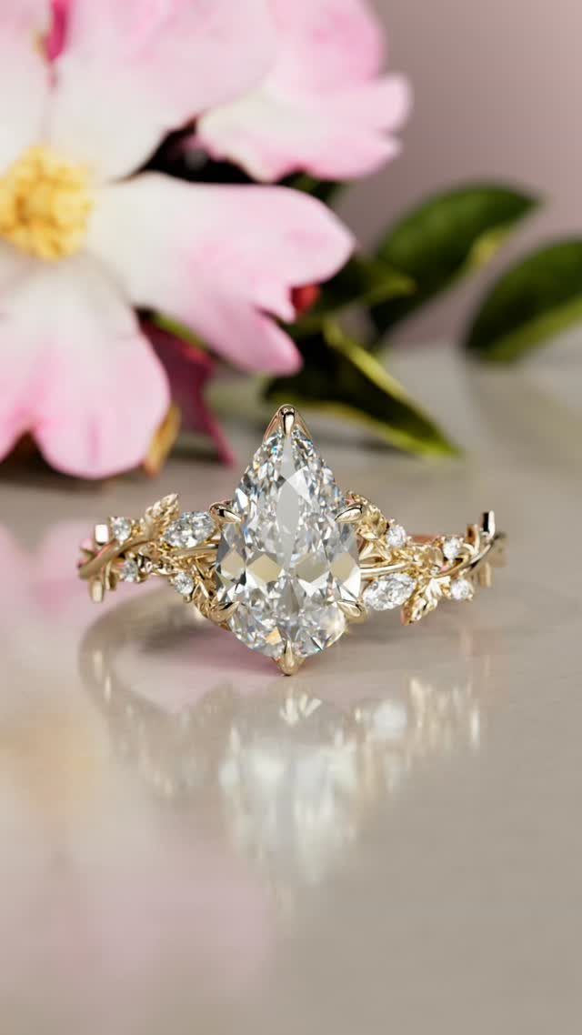 Pear-shaped diamond engagement ring with leaf-detailed gold band and pink flower background