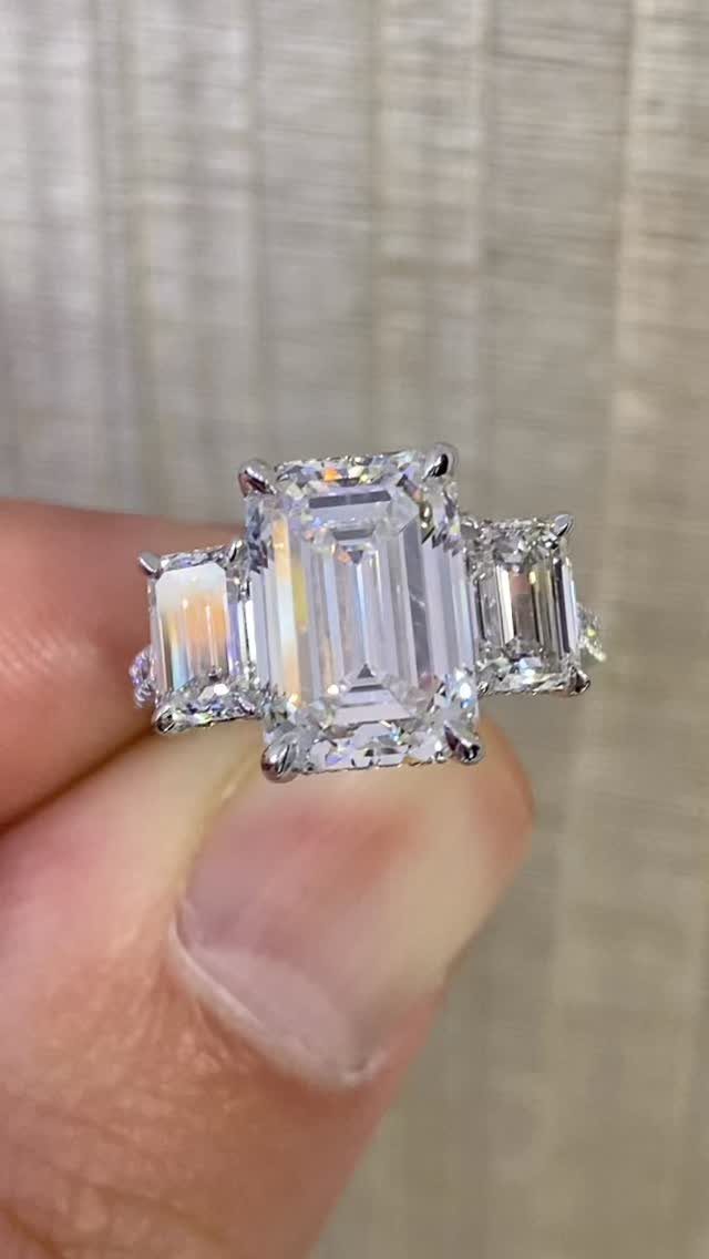 Three-stone engagement ring with emerald-cut diamonds held between a person's fingers.
