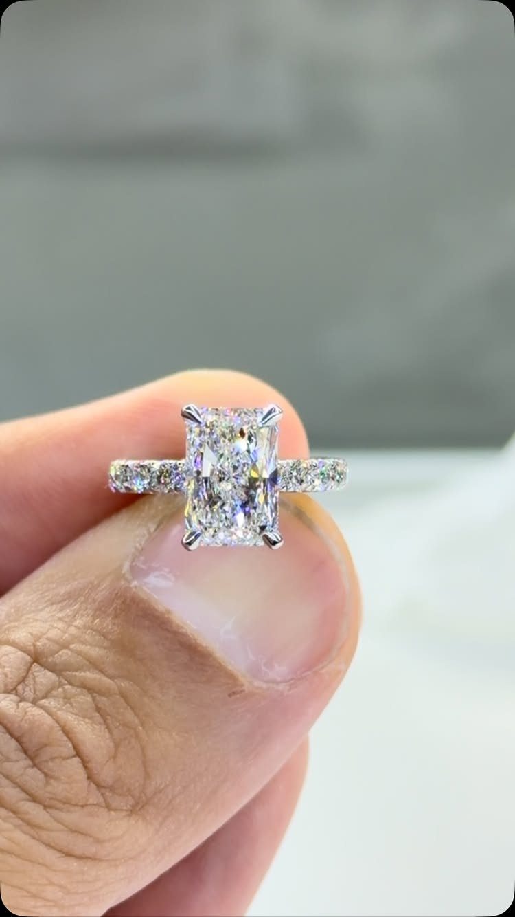 Radiant cut diamond engagement ring held between two fingers against a blurred background.