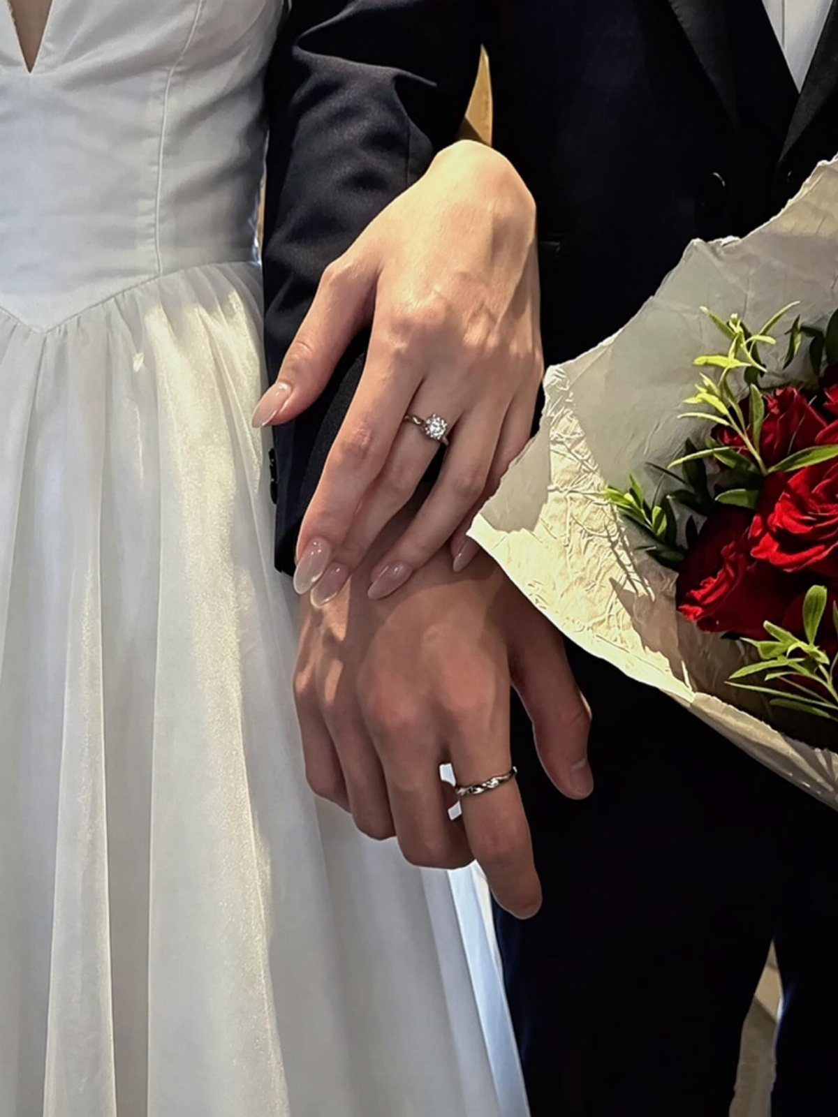 Couple's hands showing engagement and wedding rings next to a red rose bouquet