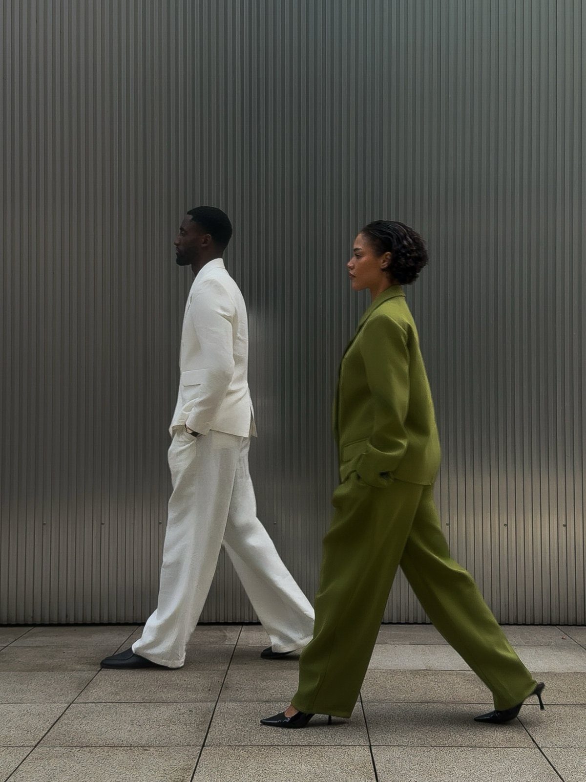 Couple in white and olive green suits walking past a grey ribbed wall.