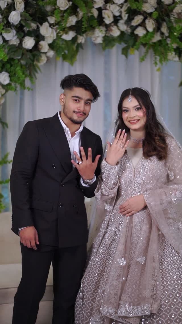 Couple posing in formal engagement attire showing their rings before a floral backdrop