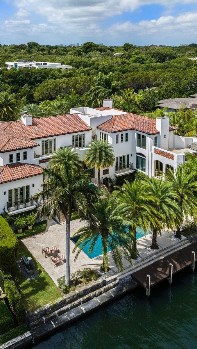 Luxury white villa with red roof, swimming pool, and palm trees by the water.