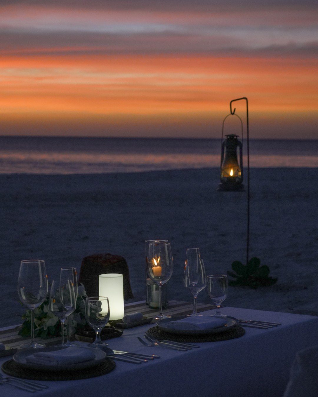 Dinner table set for two with candles on a beach at sunset.