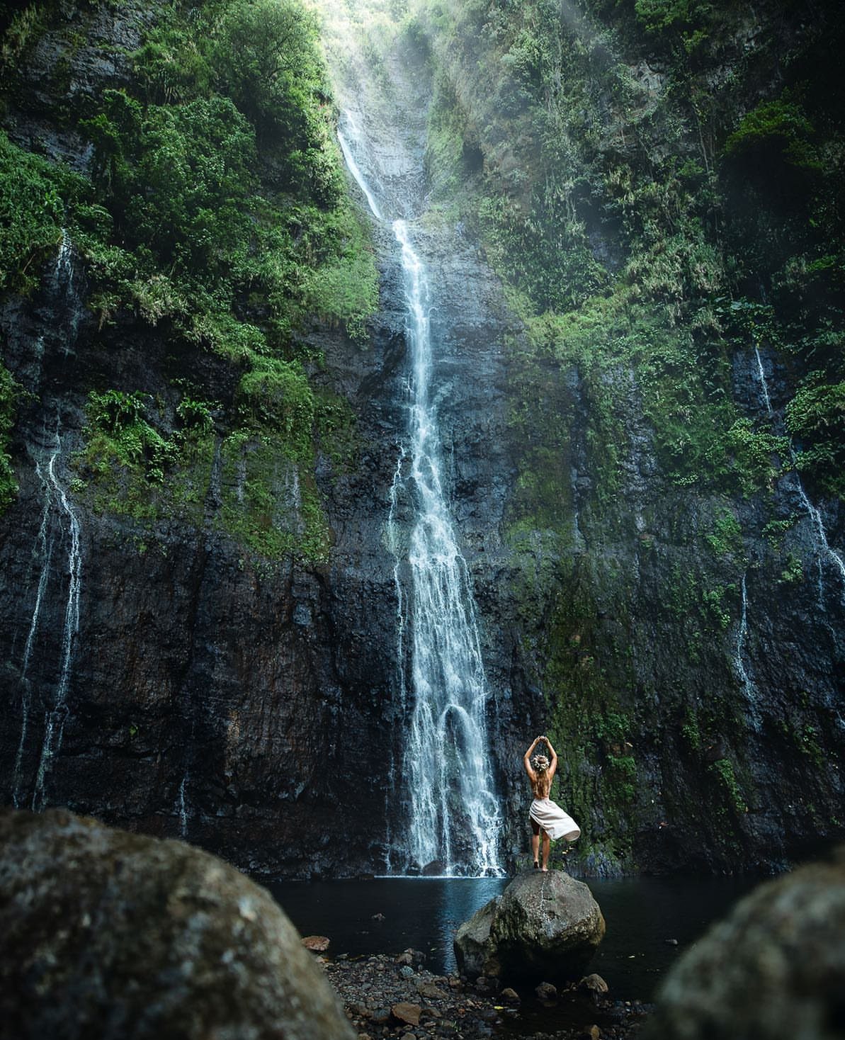 Woman in white dress posing before a tall waterfall in a lush jungle.