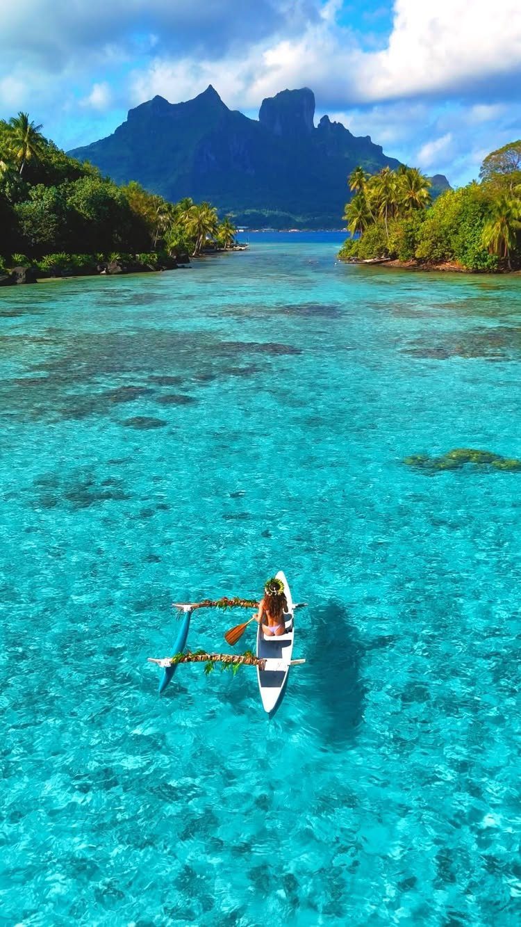 Person in a white outrigger canoe on turquoise water with tropical mountains behind.