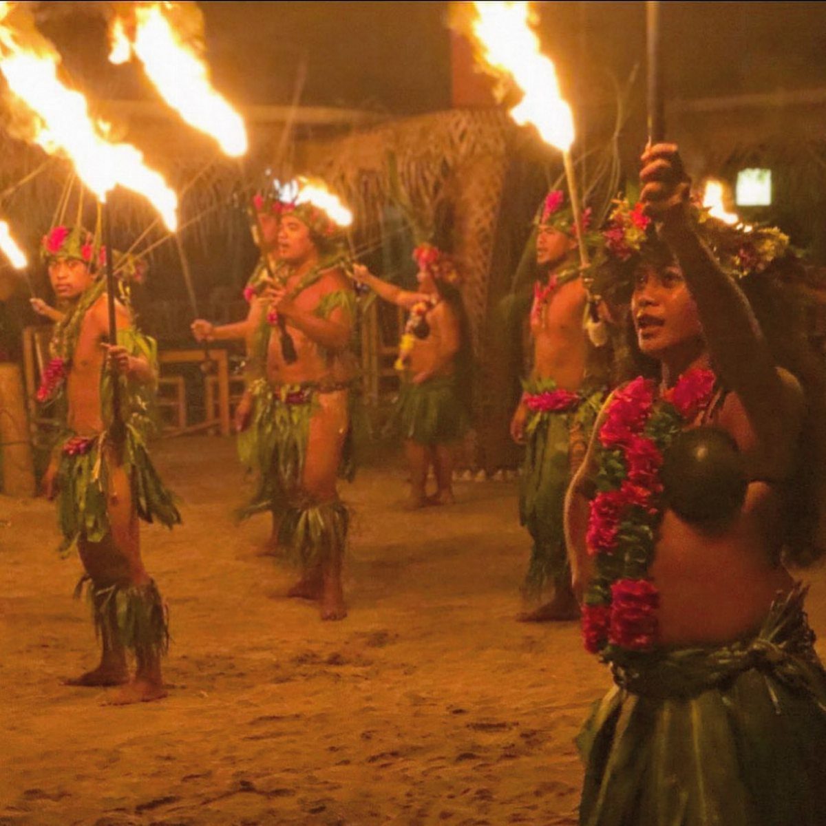 Polynesian dancers in grass skirts performing with flaming torches on a sandy beach.