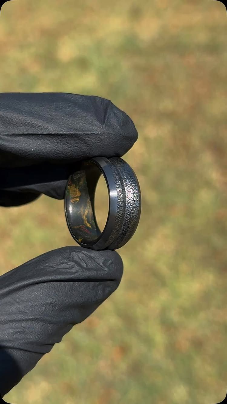 Black hammered metal wedding band held by a person wearing black gloves.