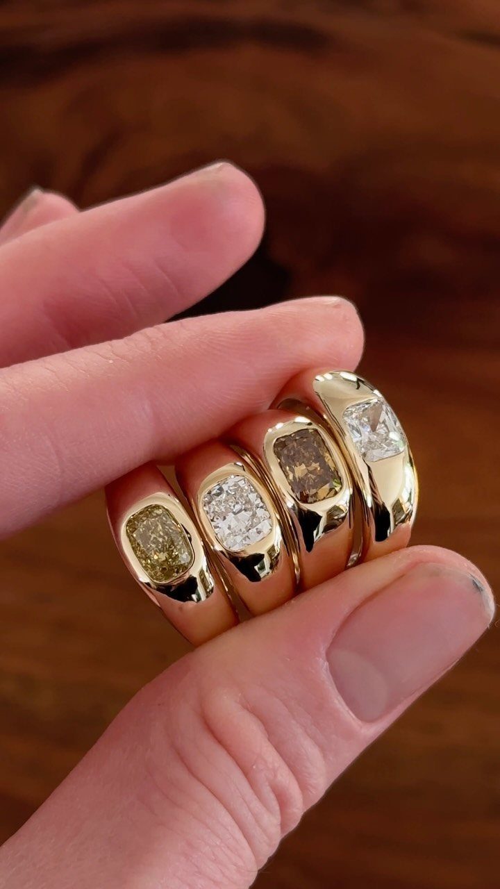 Four chunky gold rings with large colorful gemstones held between two fingers.