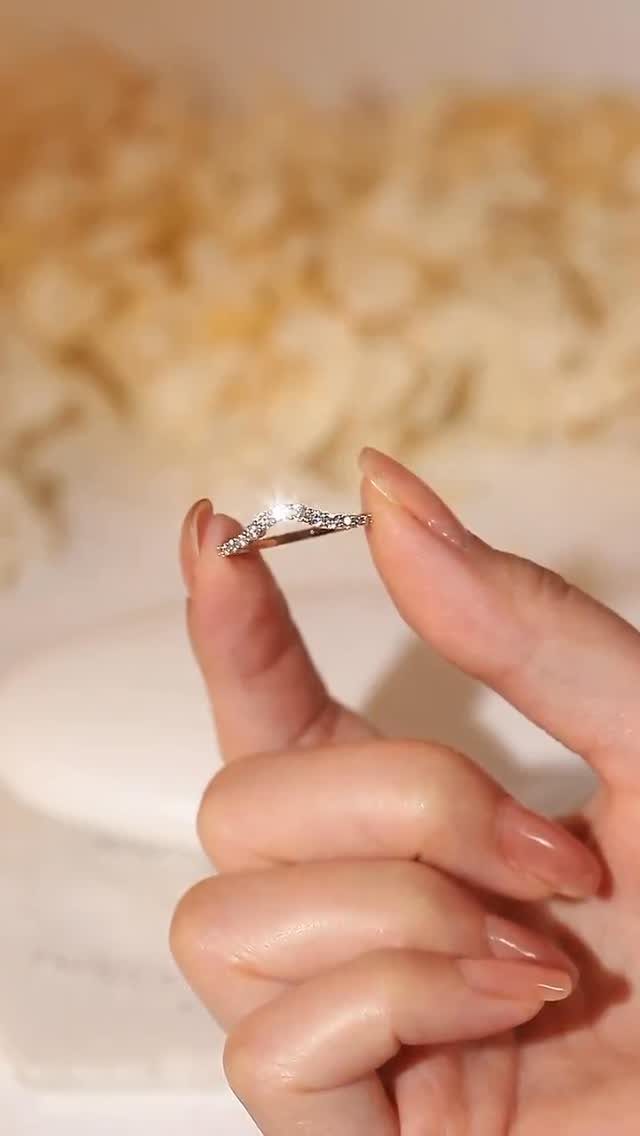 Curved diamond wedding band held between two fingers against a soft floral background.