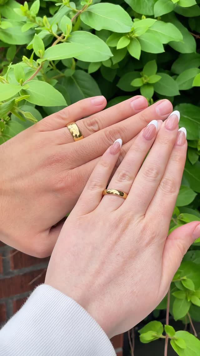 Two hands wearing gold wedding bands resting against a background of green leaves.