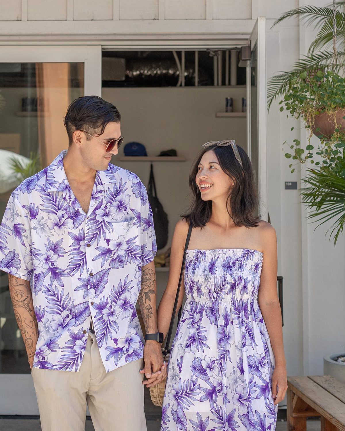 Couple wearing matching white and purple tropical floral print outfits standing outdoors.