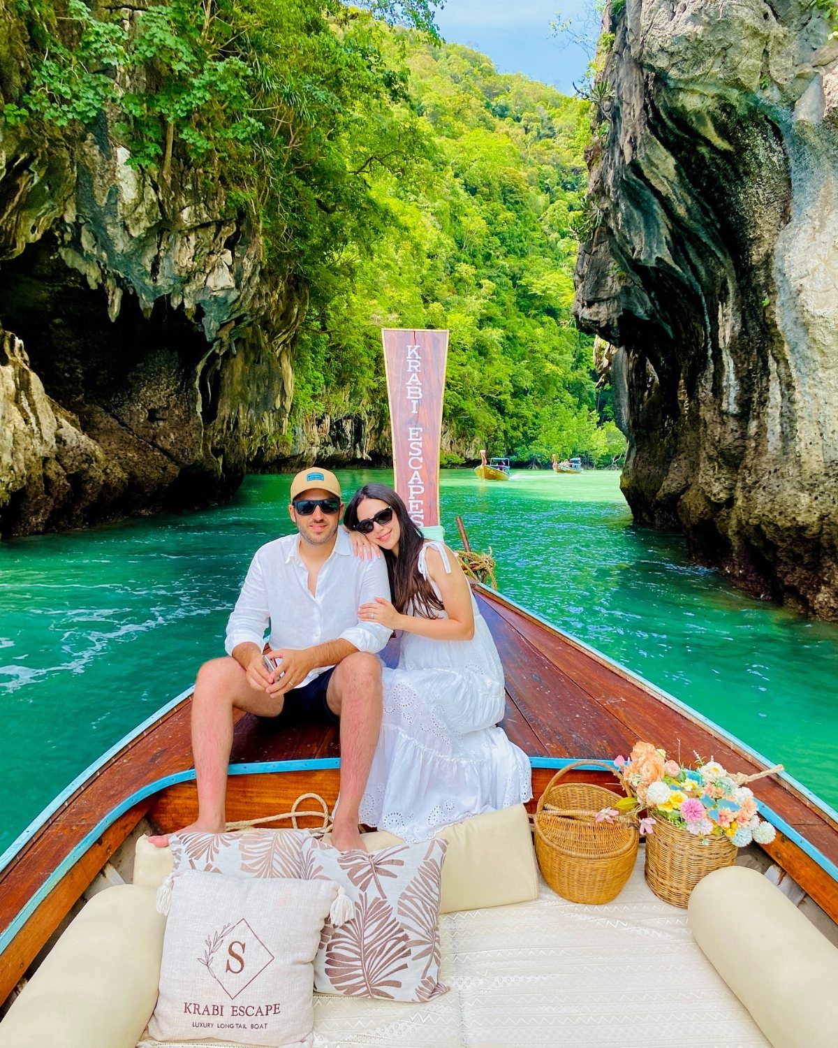 Couple in a wooden boat wearing sunglasses and white outfits on turquoise water.