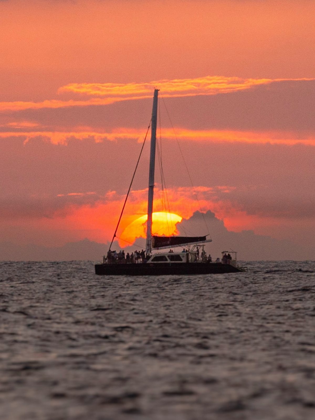 Catamaran sailboat silhouetted against a vibrant orange and red ocean sunset sky.