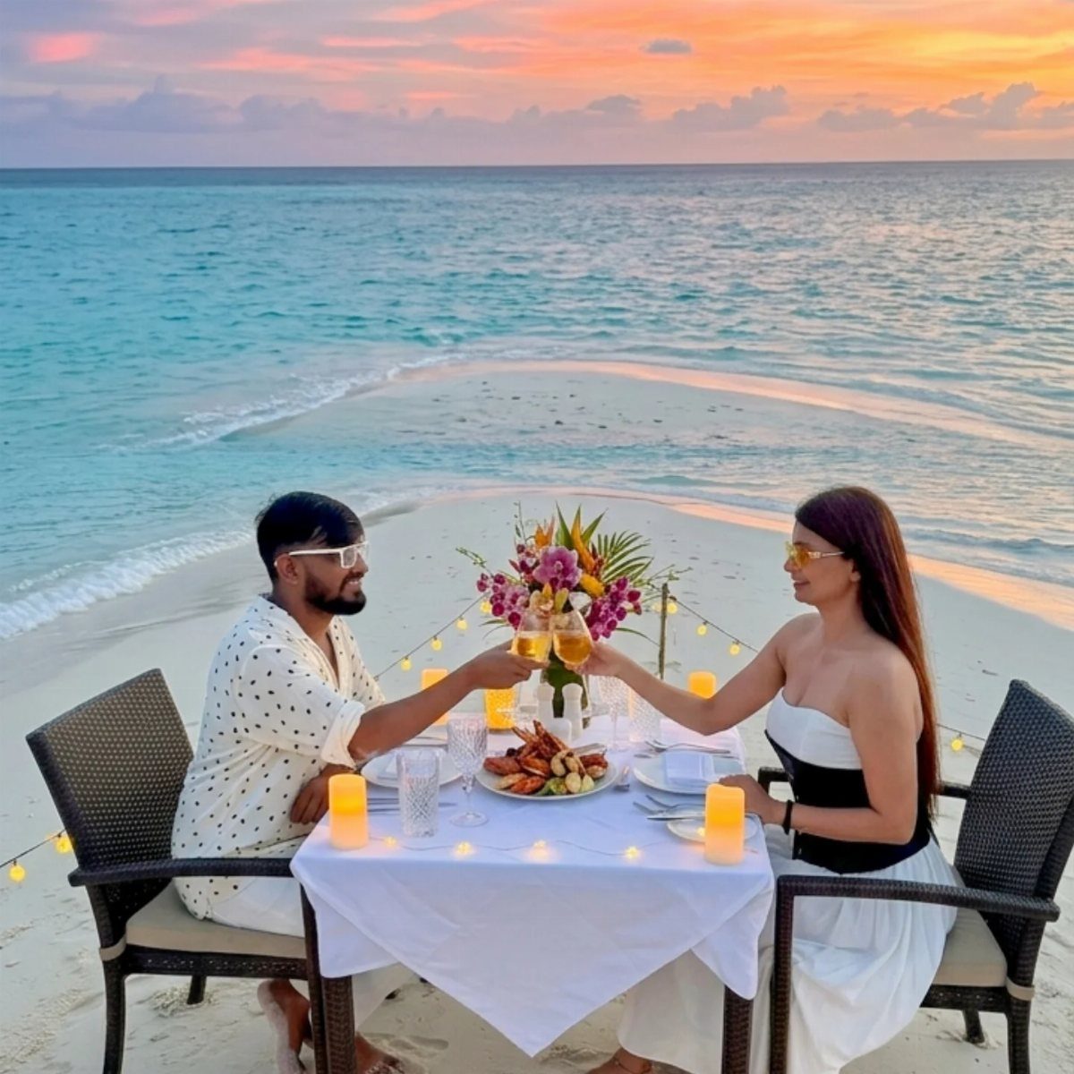 Couple clinking champagne glasses at a romantic dinner table on a sunset beach.