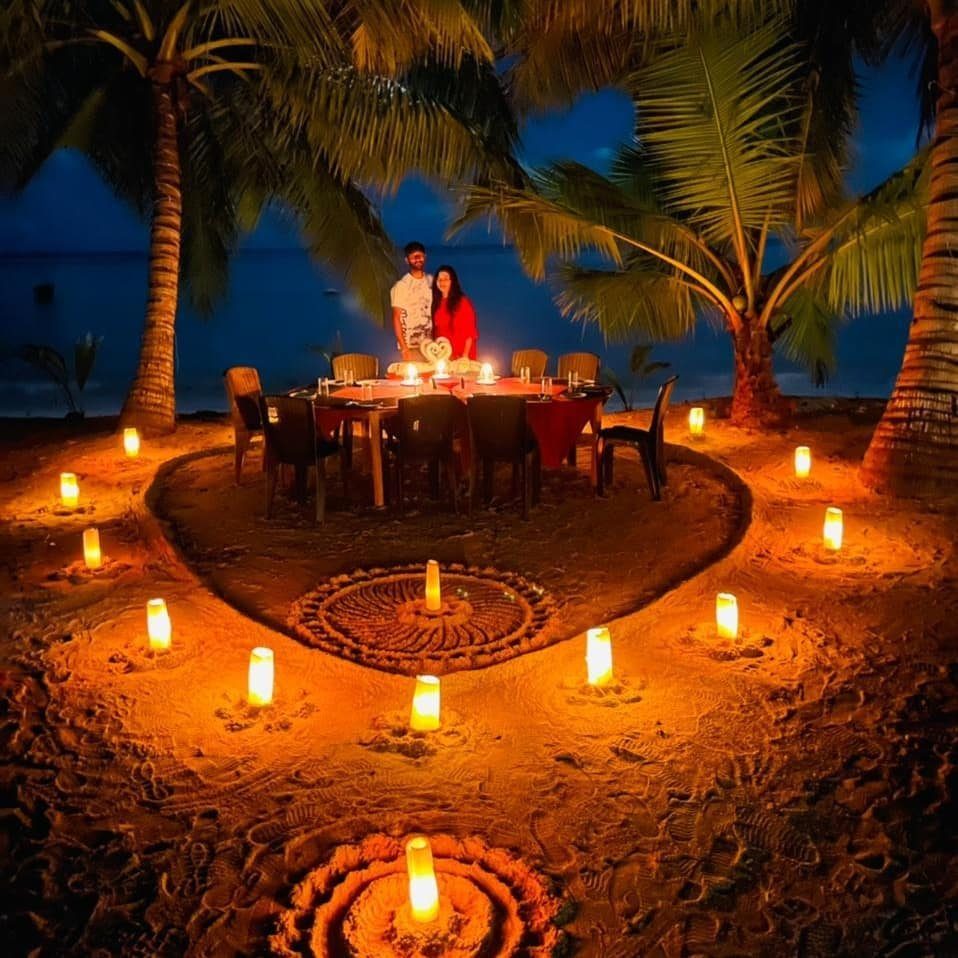 Couple standing by a candlelit dinner table on a tropical beach at dusk.