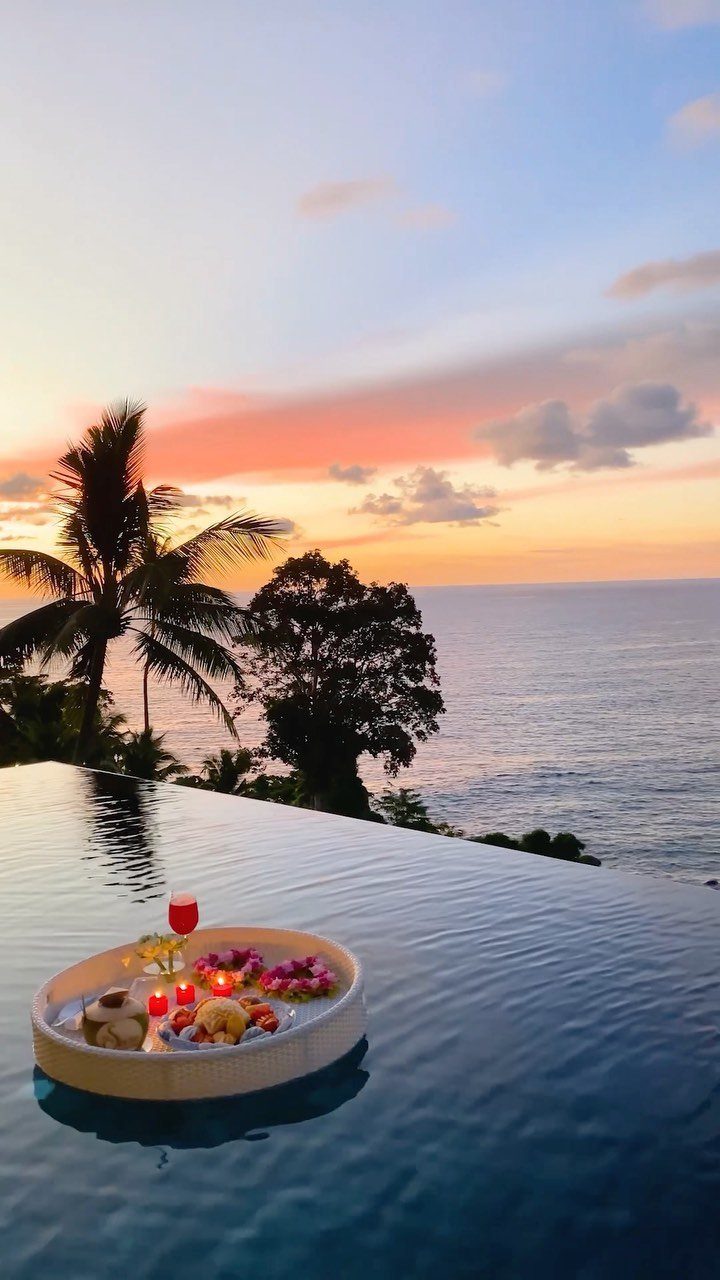 Floating breakfast tray in an infinity pool overlooking a tropical ocean sunset.