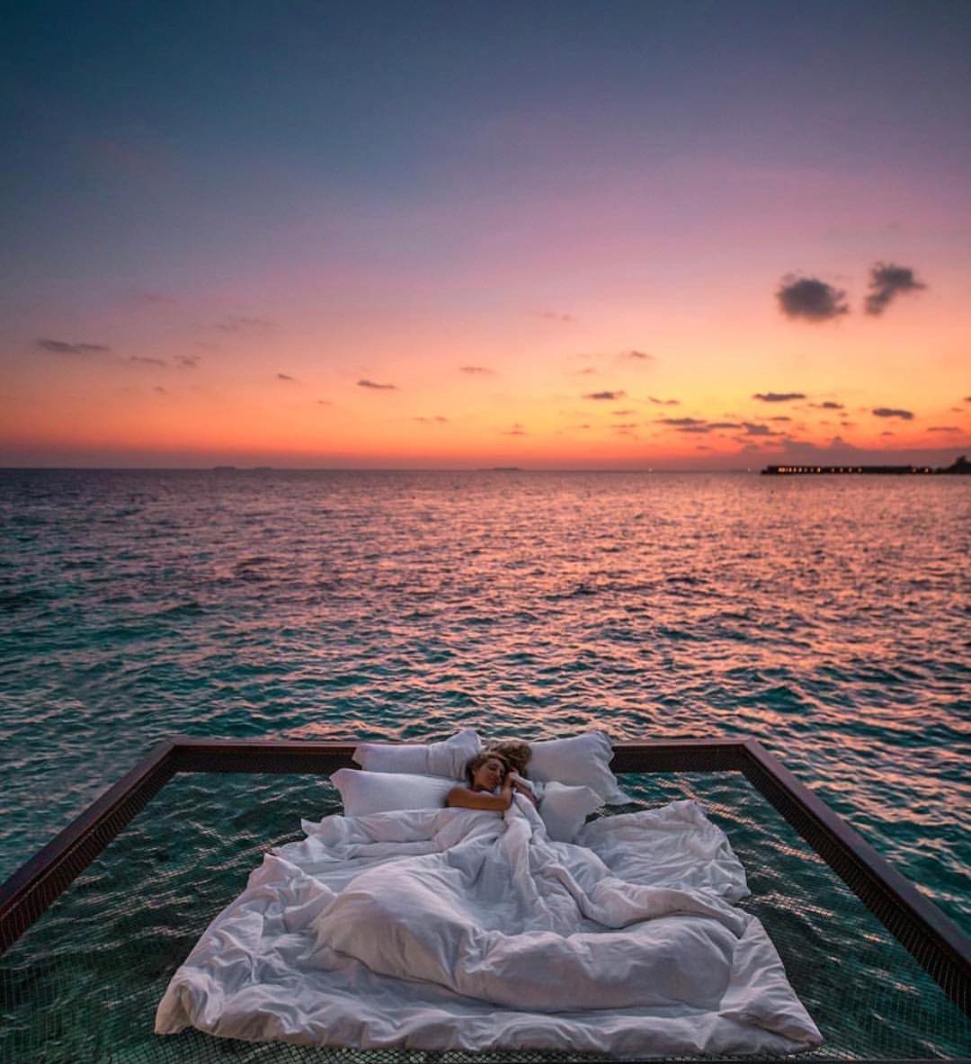 Person resting in a white bed on a glass platform over the ocean at sunset.