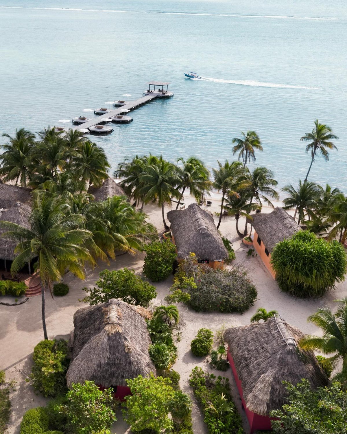 Tropical beach resort with thatched-roof bungalows, palm trees, and a long wooden pier.