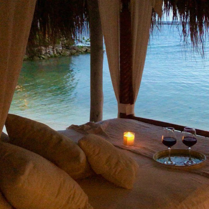 Romantic bed setup with wine and candles overlooking a turquoise tropical beach resort.