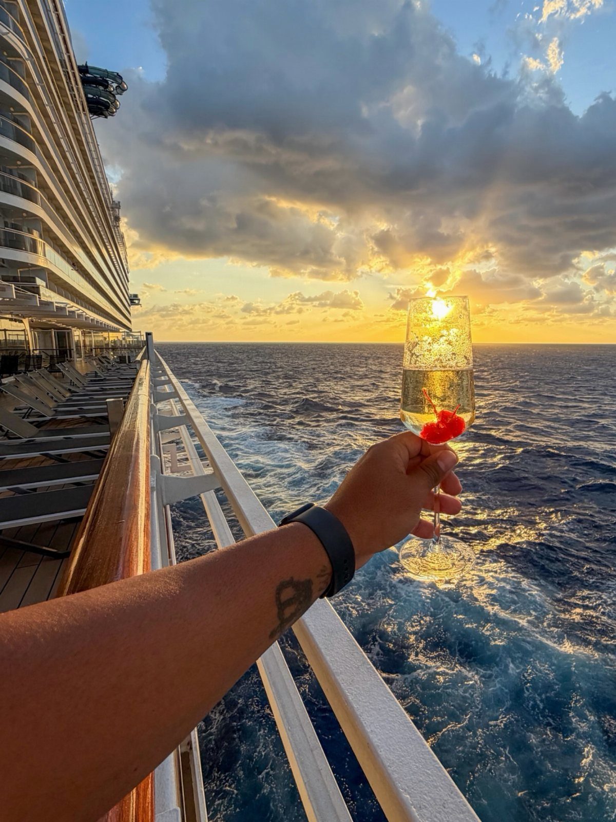 Champagne glass held against a golden sunset over the ocean from a cruise ship deck.