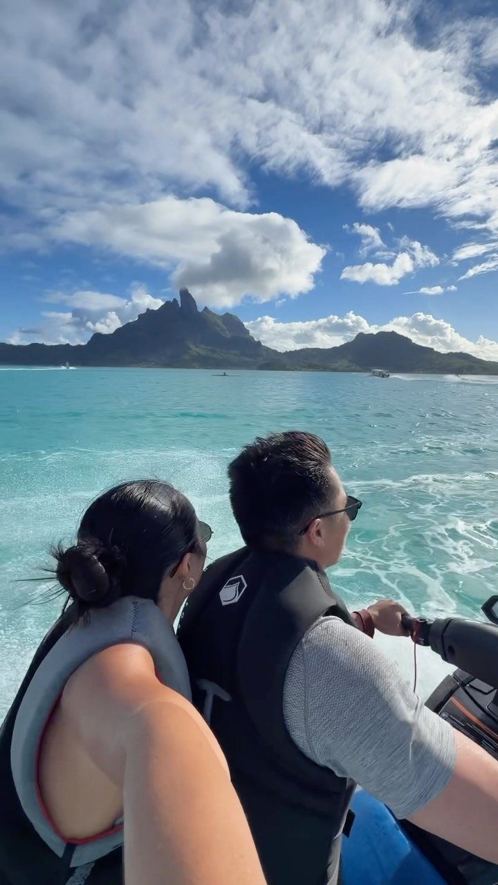 Couple on a blue jet ski riding through turquoise water toward tropical mountains.