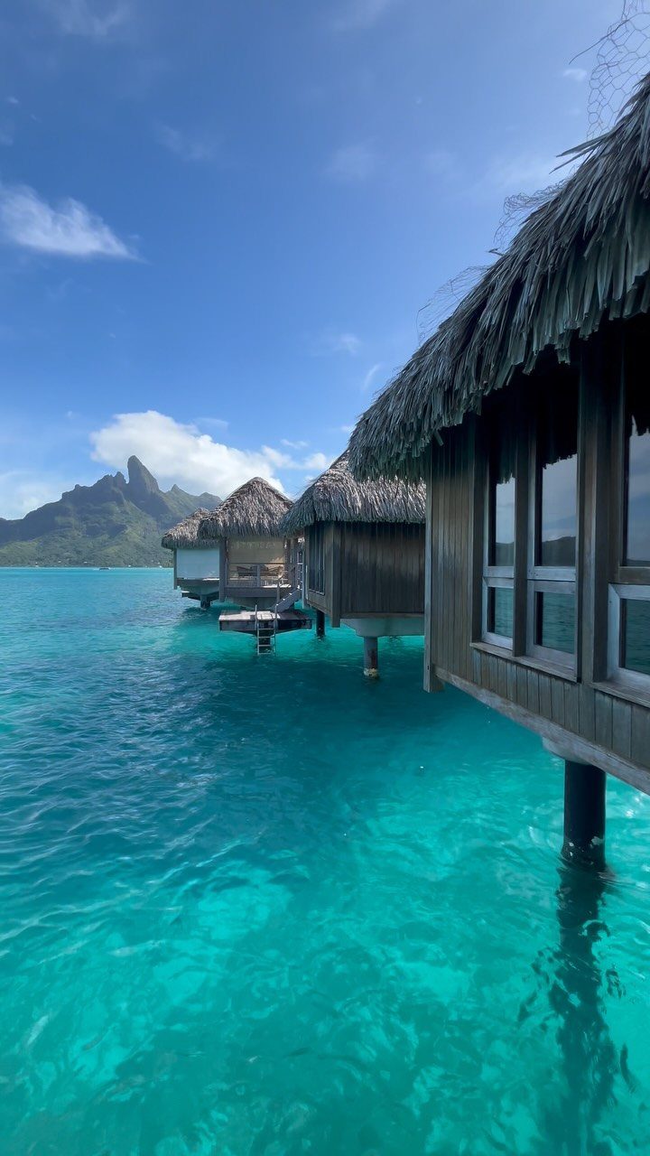 Overwater bungalows with thatched roofs over turquoise water with a mountain in background