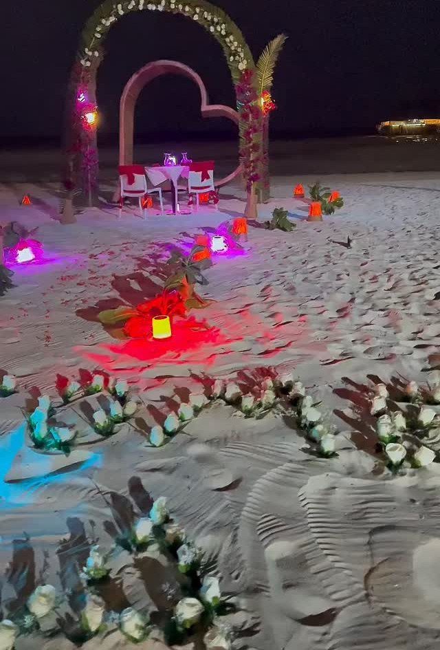 Romantic beach dinner setup with a heart backdrop, floral arch, and glowing lanterns.