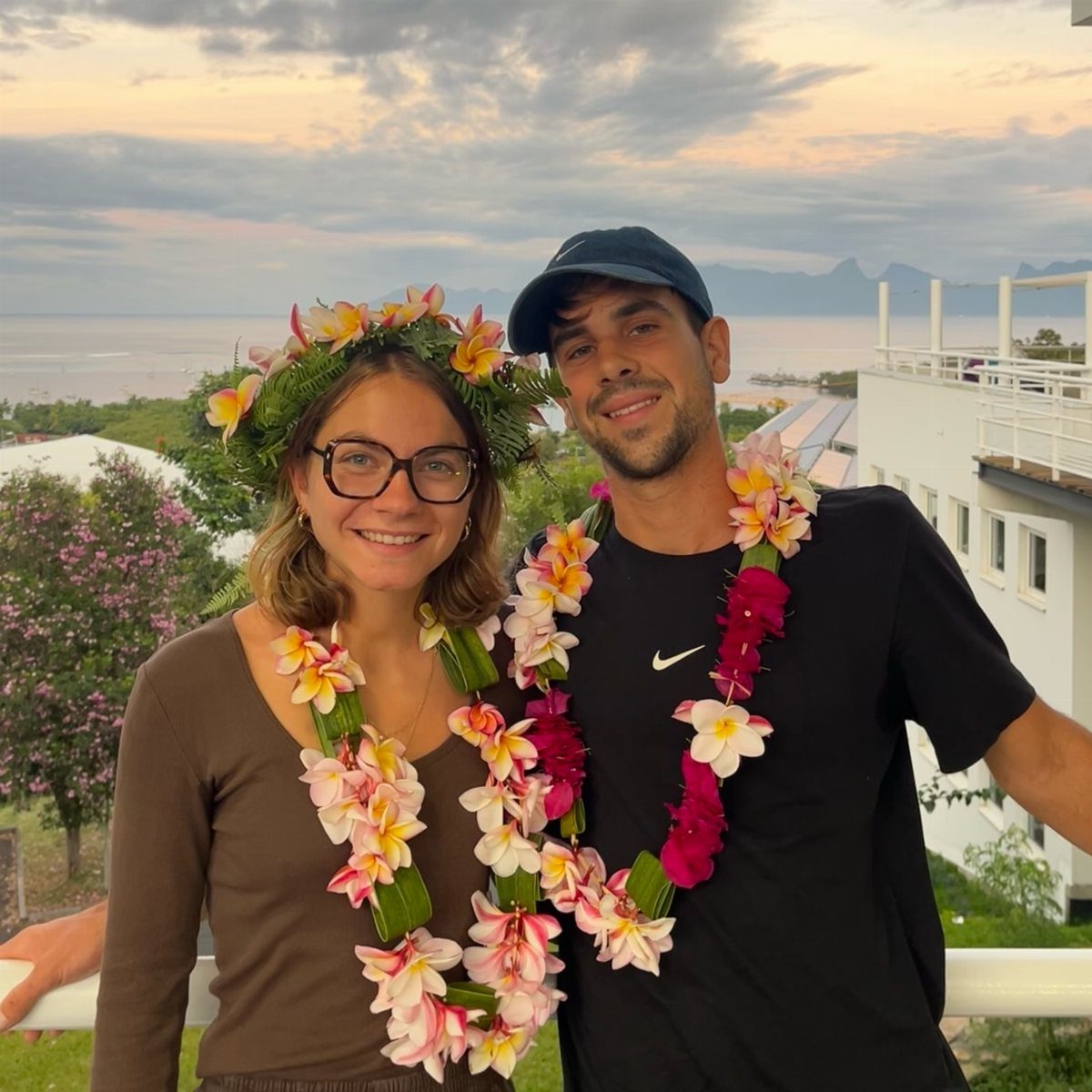 Couple wearing Tahitian flower garlands and crown posing against a tropical coastal backdrop.