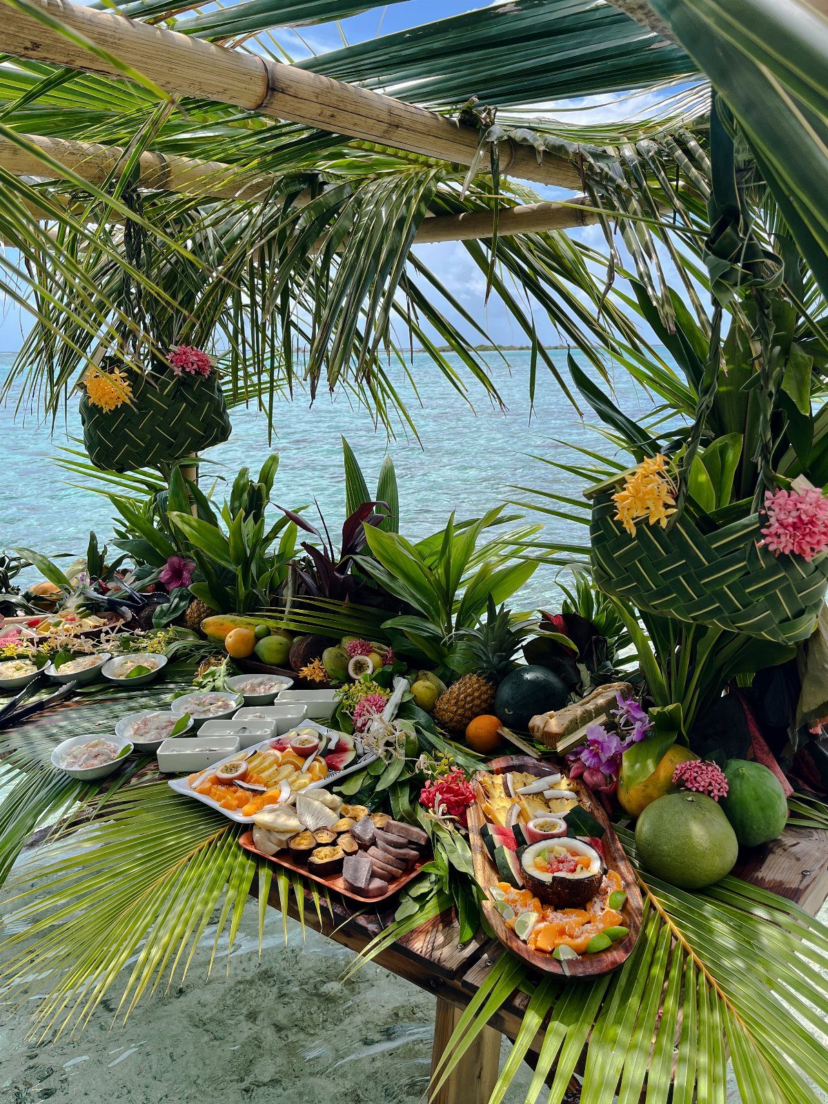 Tropical food spread with fresh fruit and seafood under a palm leaf canopy.