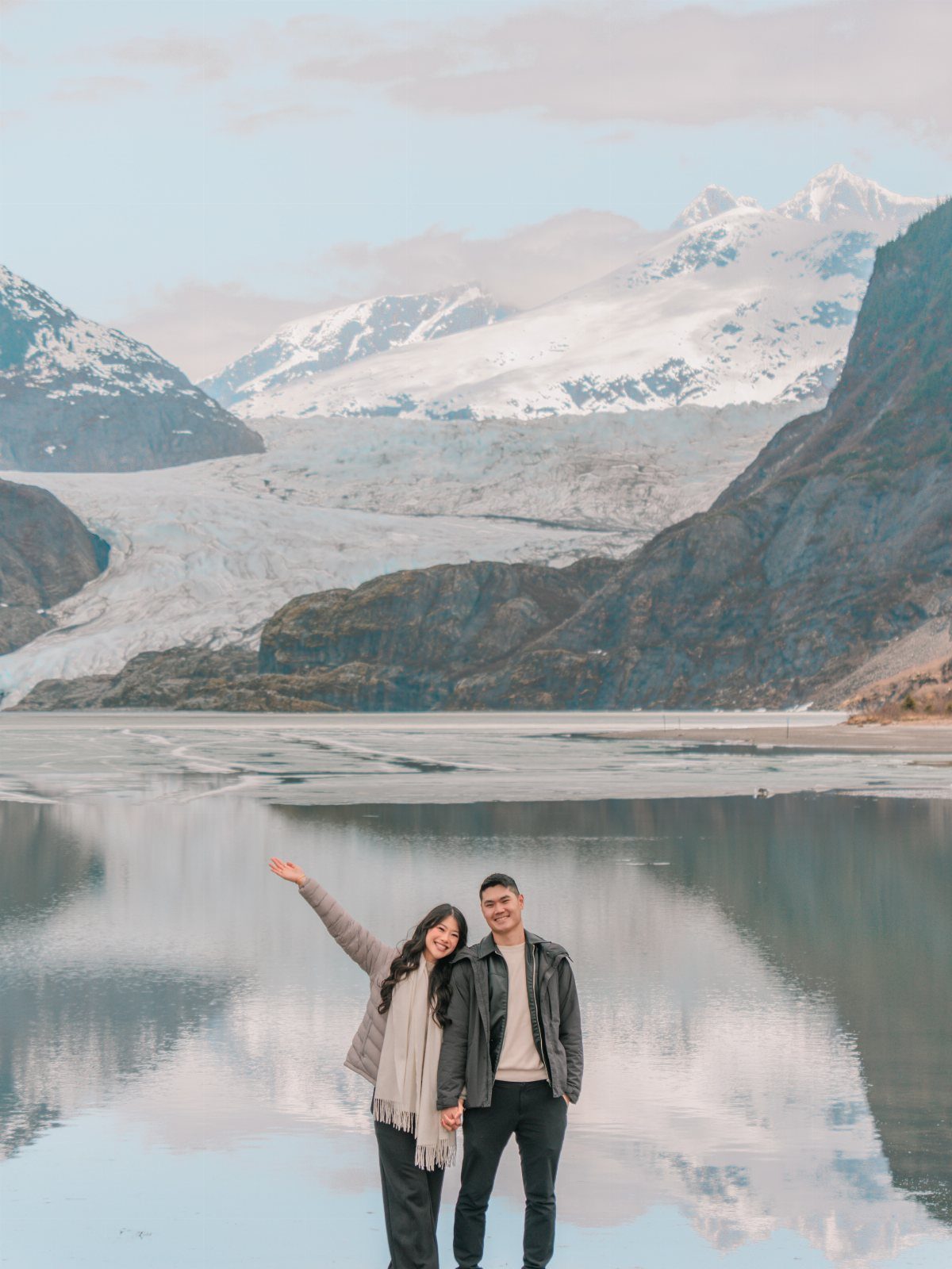 Couple standing by a reflective lake with a massive glacier and snowy mountains.