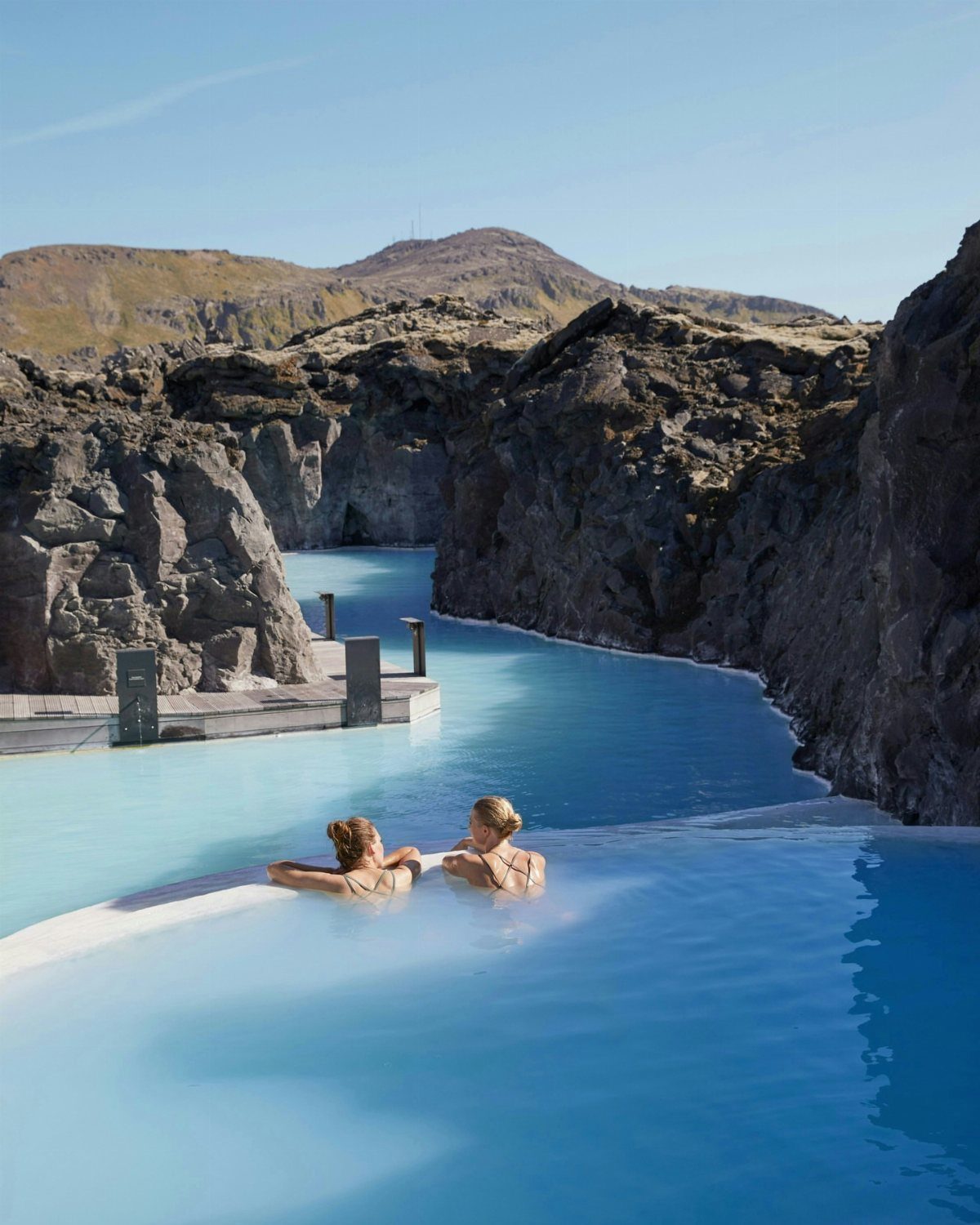 Two people relaxing in a bright blue geothermal hot spring among volcanic rocks.