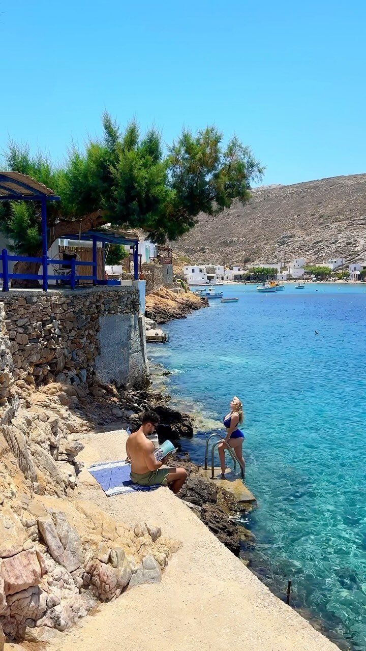 Couple relaxing by clear turquoise water along a rocky Greek coastline with white buildings.