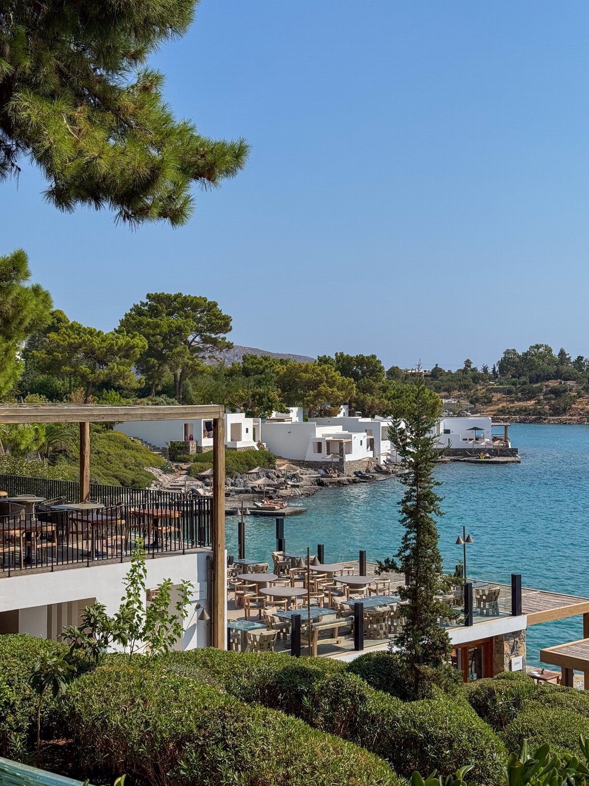 White waterfront bungalows and outdoor dining terrace overlooking a turquoise Greek bay.