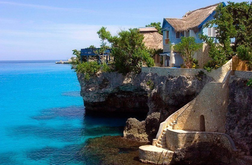 Cliffside hotel with thatched roofs and stone stairs overlooking turquoise Jamaican ocean waters.