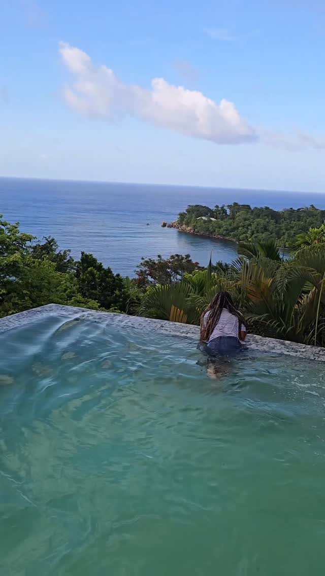 Person in an infinity pool overlooking a tropical coastline and blue ocean.