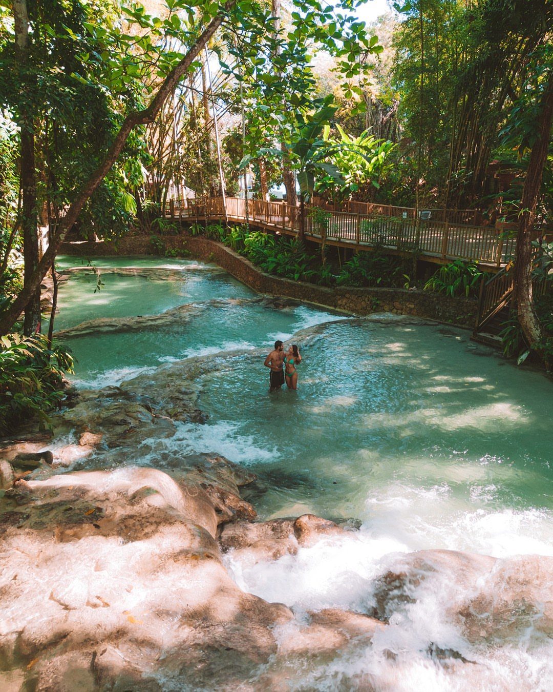 Couple standing in turquoise natural spring pools surrounded by lush tropical greenery.