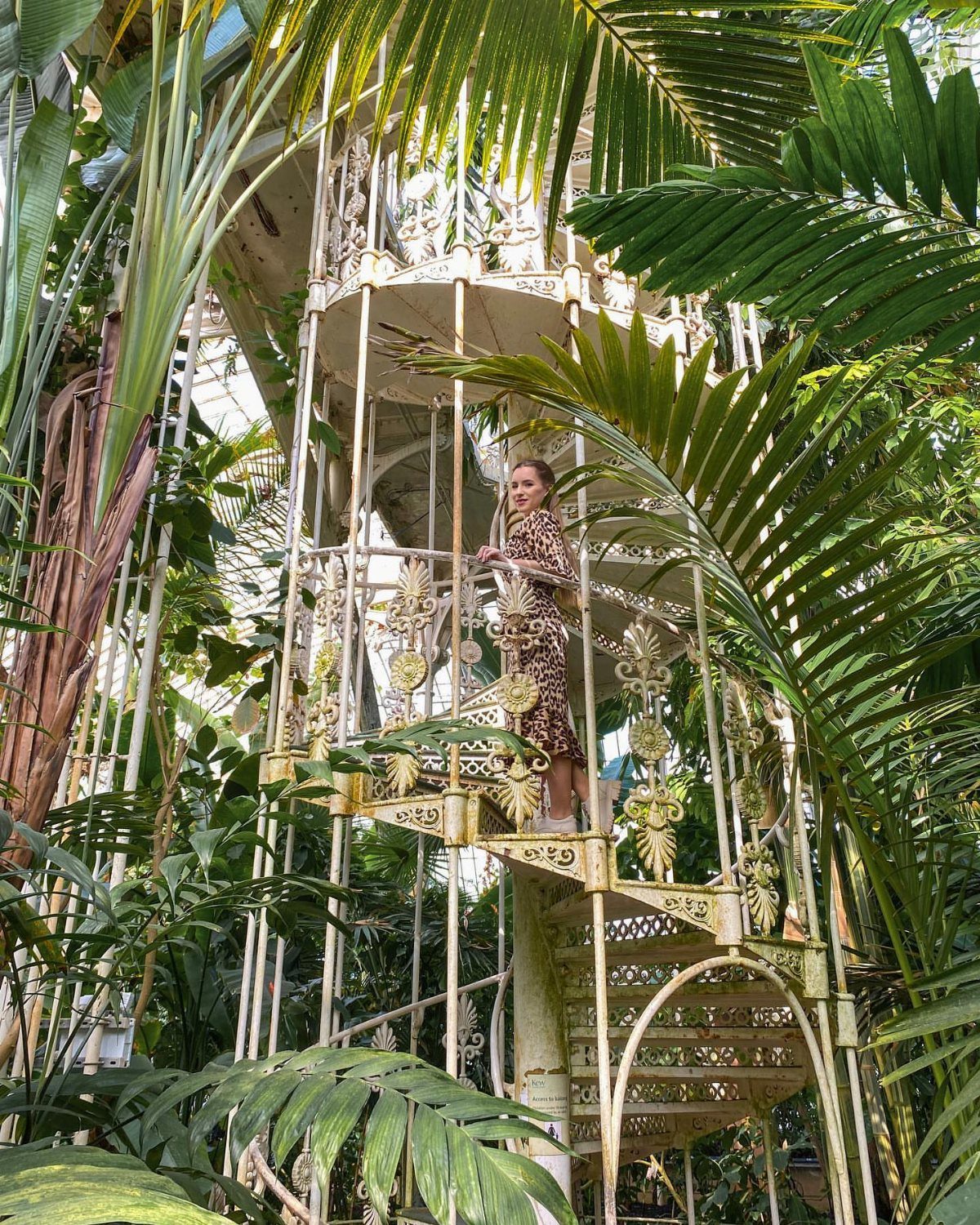 Woman in patterned dress standing on ornate white spiral staircase in tropical conservatory