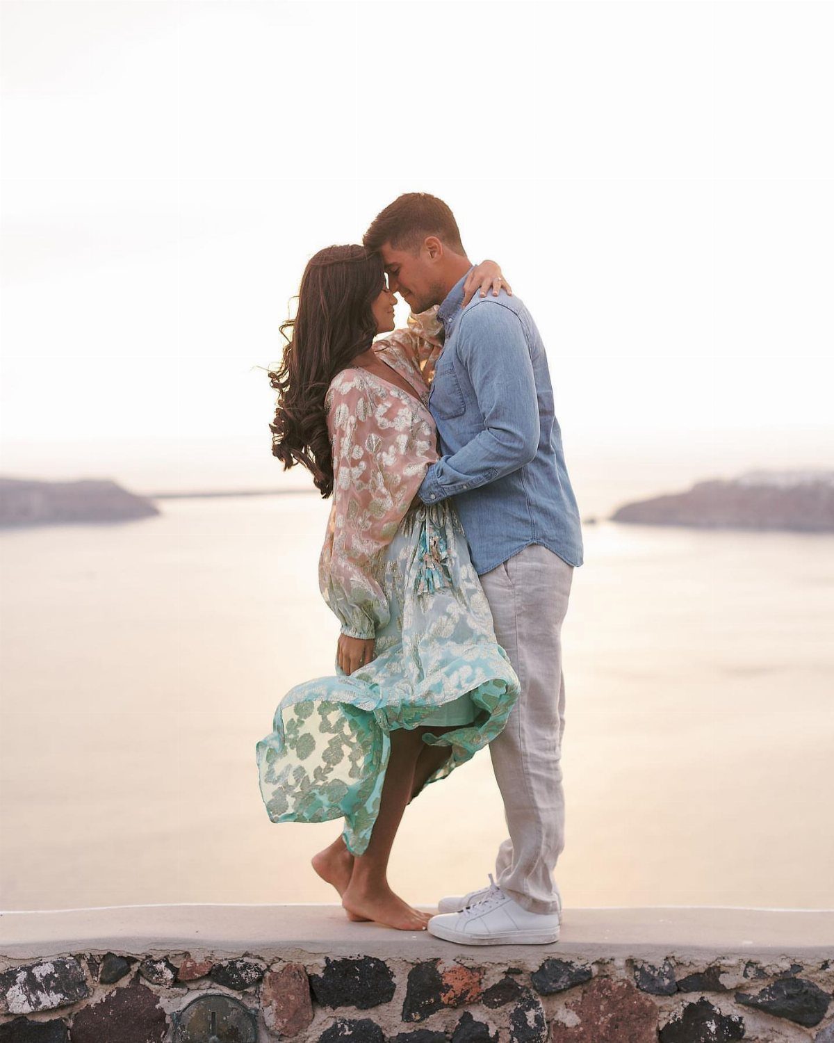 Couple embracing on a stone ledge overlooking the sea in Santorini at dusk.